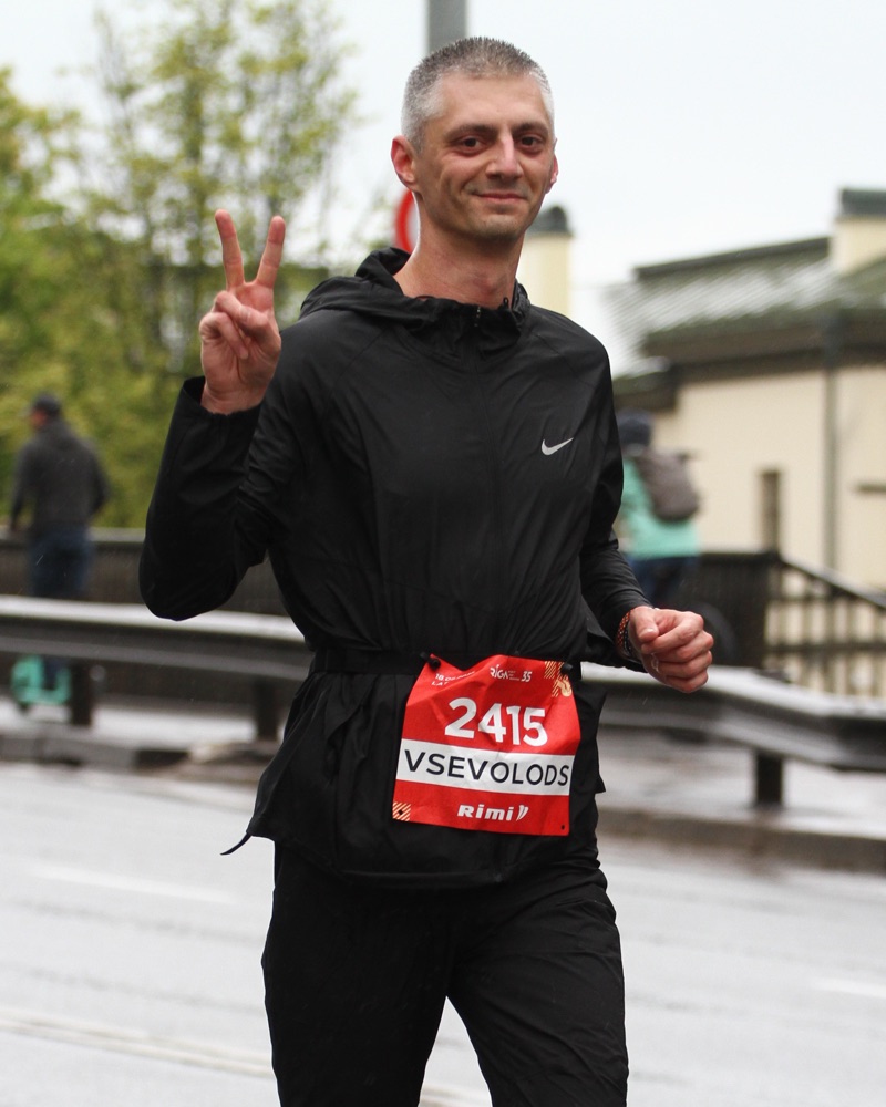 Vsevolod mid-race on a wet road in a black running kit with bib 2415, making a peace sign