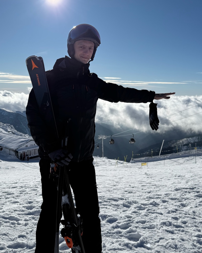 Vsevolod on a ski slope in a black jacket and helmet with skis on his shoulder, and a sea of cloud and a gondola line behind him